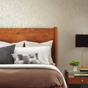 Bedroom - A cozy features a wooden headboard, layered pillows, and a brown comforter. A sleek black lamp and small potted plant rest on the bedside table. The walls boast York Wallcoverings' Neutral & Silver Nazca Wallpaper, adding a textured vertical pattern that enhances the modern design