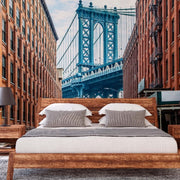 Bedroom - A modern bedroom with a large bed centered between two nightstands, each with a lamp, against a vivid New York Bridge Wallpaper Mural from Decor2Go Wallpaper Mural featuring the Manhattan Bridge and surrounding brick buildings.