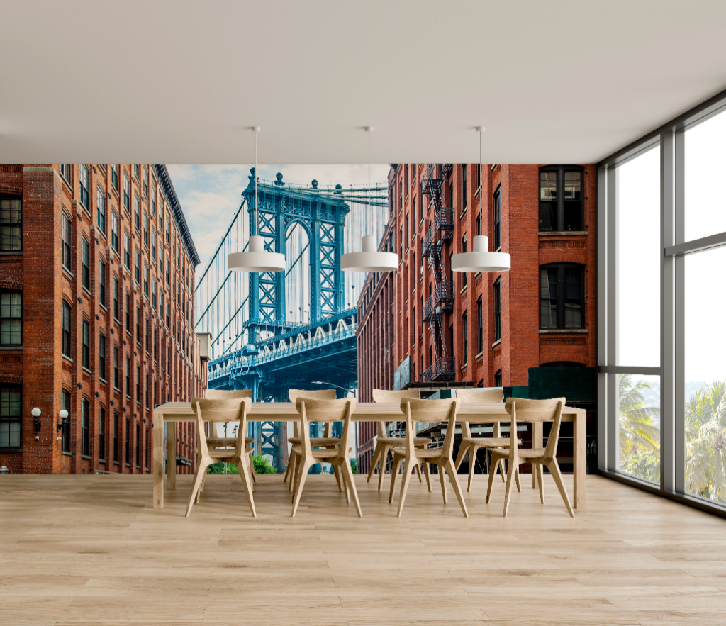 Dining Room - A spacious dining room features a large table with chairs, set against a wall mural of the Brooklyn Bridge flanked by historic red brick buildings showcasing New York landmarks. Large windows offer a view of green trees featuring the Decor2Go Wallpaper Mural New York Bridge Wallpaper Mural.