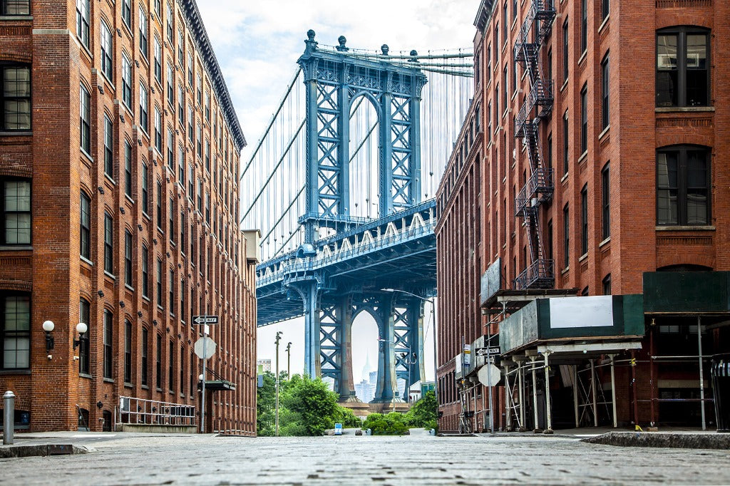 View of the Manhattan Bridge framed by red brick buildings on a cobblestone street, with a clear blue sky in the background, showcasing Decor2Go Wallpaper Mural's New York Bridge Wallpaper Mural's architectural design.