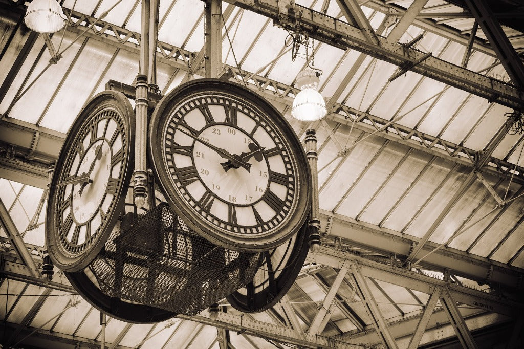 Vintage double-faced hanging On time Clockwork Wallpaper Mural in a railway station, featuring roman numerals and set against an old, iron-beamed roof. Monochrome, sepia-toned image enhancing the historic ambiance of this clock by Decor2Go Wallpaper Mural.
