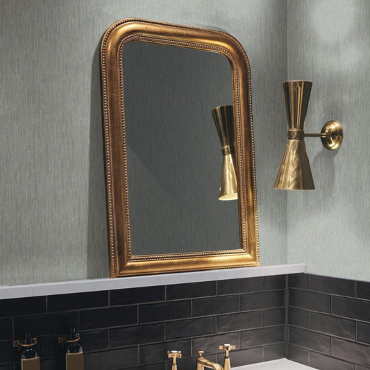 A bathroom with York Wallcoverings' Pale Green & Silver Metallic Cascade Wallpaper features a gold-framed mirror, accented by a brass wall sconce. Below, the black tiled backsplash and white countertop highlight black and gold faucets for a sleek metallic effect.