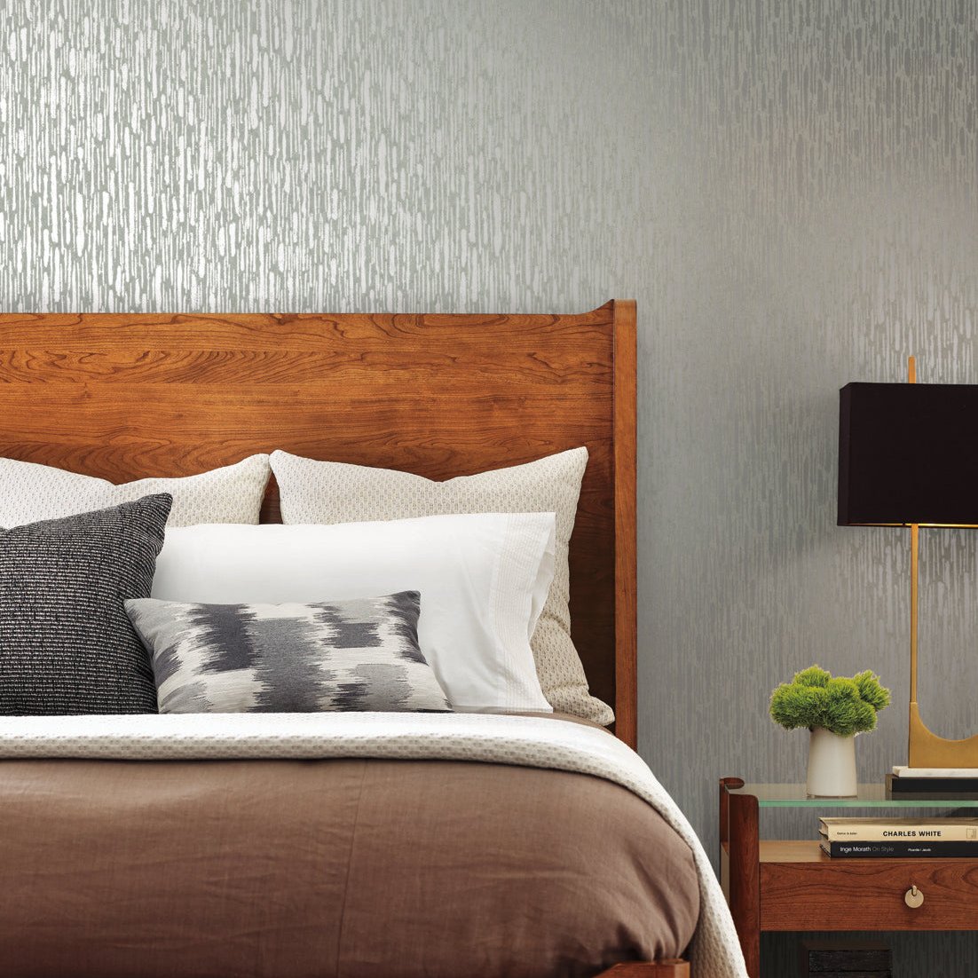 A cozy bedroom features a wooden headboard with white and gray pillows, brown bedding, and a nightstand holding a small green plant and black lamp. The wall is adorned with York Wallcoverings' Pale Green & Silver Metallic Cascade Wallpaper, adding sophistication to the space.