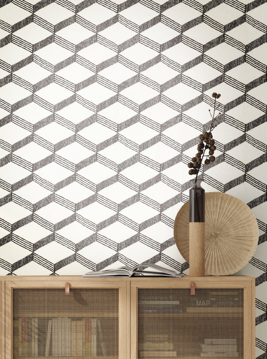 Displayed above a wooden cabinet, the York Wallcoverings Palisades Paperweave Wallpaper in Beige/Black, featuring a geometric 3D cube pattern, brings a touch of timeless design to the space. The cabinet is filled with books and showcases a round decorative object along with a tall vase containing dried branches, enhancing the scene with natural texture.