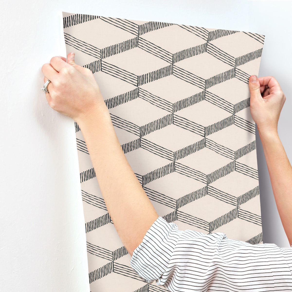 While applying the York Wallcoverings' Palisades Paperweave Wallpaper in Beige/Black, featuring a black and white geometric cube pattern, the person's rolled-up sleeves partly expose their striped shirt. This classic design adds an elegant touch to the white wall.