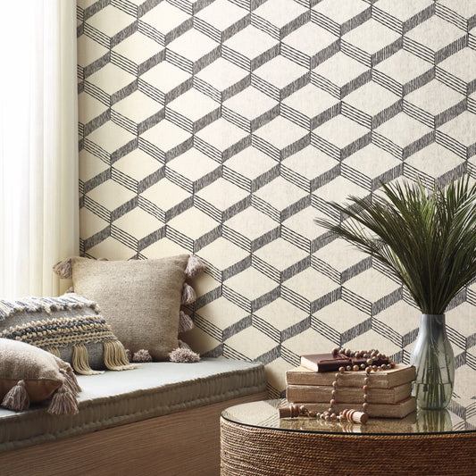 A cozy seating area features a cushioned bench adorned with tassel pillows. The wall is beautifully covered with York Wallcoverings' Palisades Paperweave Wallpaper in Beige/Black, which adds a touch of natural texture. A round glass table holds a stack of books and a potted plant, as sunlight filters through a sheer curtain.