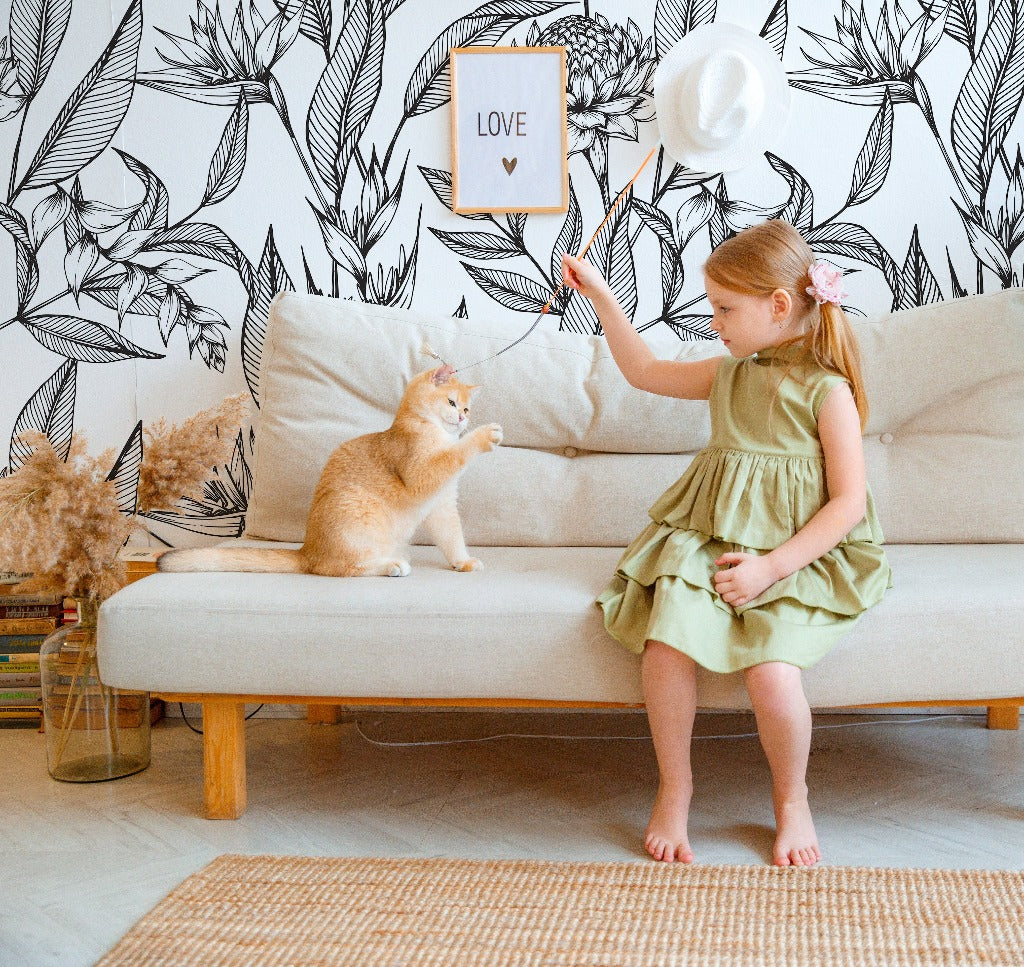 A young girl in a green dress plays with a ginger cat on a beige sofa, against Decor2Go Wallpaper Mural's Paradise Flower Wallpaper Mural background. Nearby, a soft toy and a hat decorate the area.