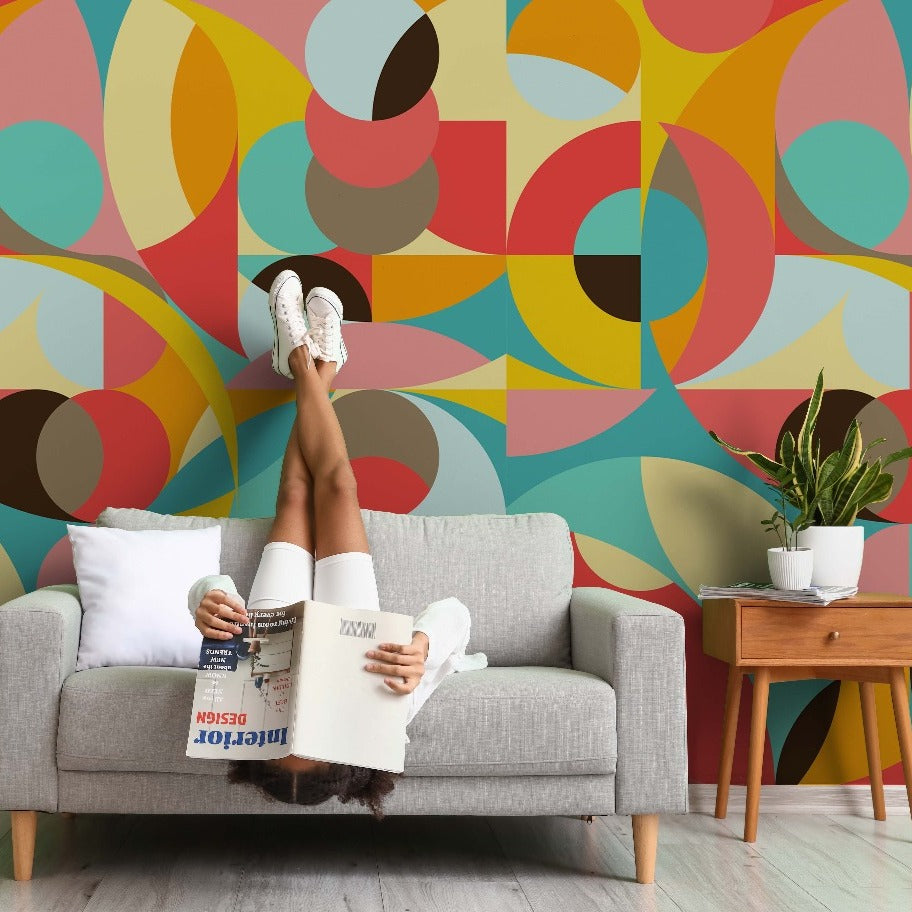 A person lying upside down on a gray sofa, reading a magazine, with their legs up against a vibrant, Decor2Go Wallpaper Mural, Pastel Jungle Wallpaper Mural. A side table with a plant is next to the sofa.
