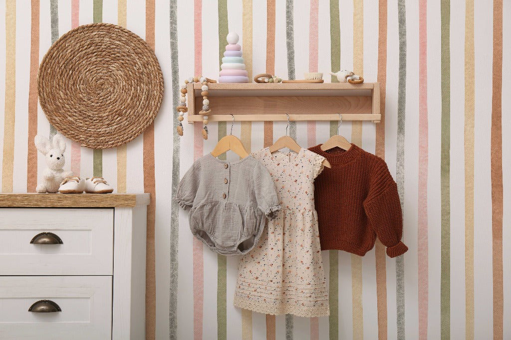 A cozy children's room wall with Pastel Perfection Wallpaper Mural from Decor2Go Wallpaper Mural, decorated with a hanging wicker plate, a wooden shelf with toys, and clothes on hooks below it. A dresser is to the