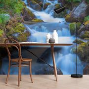 Home Office - A serene home office setup with a wooden chair and desk against a large, vivid nature mural of a Decor2Go Wallpaper Mural Peaceful Waterfall Wallpaper Mural surrounded by mossy rocks, complemented by a black desk lamp and white decorative vase.
