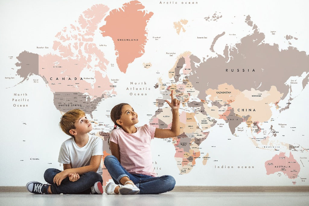 Two children sitting on the floor, pointing at a Decor2Go Peach World Map Wallpaper Mural on the wall, engaged in learning about different countries.