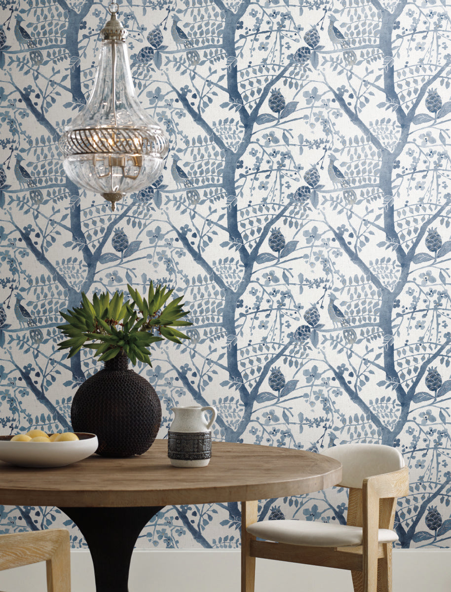 A round wooden table adorned with a black vase containing green foliage, a small pitcher, and a bowl of lemons sits under a modern glass chandelier. The background showcases Peacock Block Print Wallpaper in blue by York Wallcoverings, which features blue and white botanical patterns with branches and leaves designed from durable materials for a nature-inspired aesthetic.