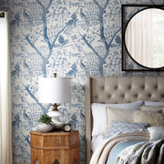 Bedroom - A cozy showcasing a bed with white and beige bedding, a tufted headboard, and a round mirror on the wall. A wooden nightstand supports a white lamp, a plant, and a clock. The York Wallcoverings Peacock Block Print Wallpaper in Blue (60 SqFt) adds an elegant touch to the nature-inspired design