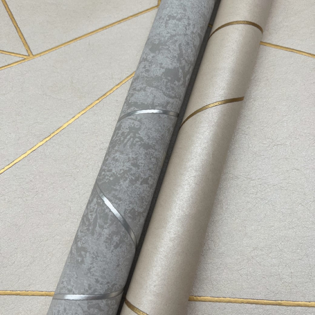 Two rolls of York Wallcoverings lean against a wall with geometric patterns. One roll, Pearl & Gold Nazca Wallpaper (57 SqFt), is beige accented in gold metallic finishes, while the other is gray with silver. The background features cream sections outlined in gold lines.