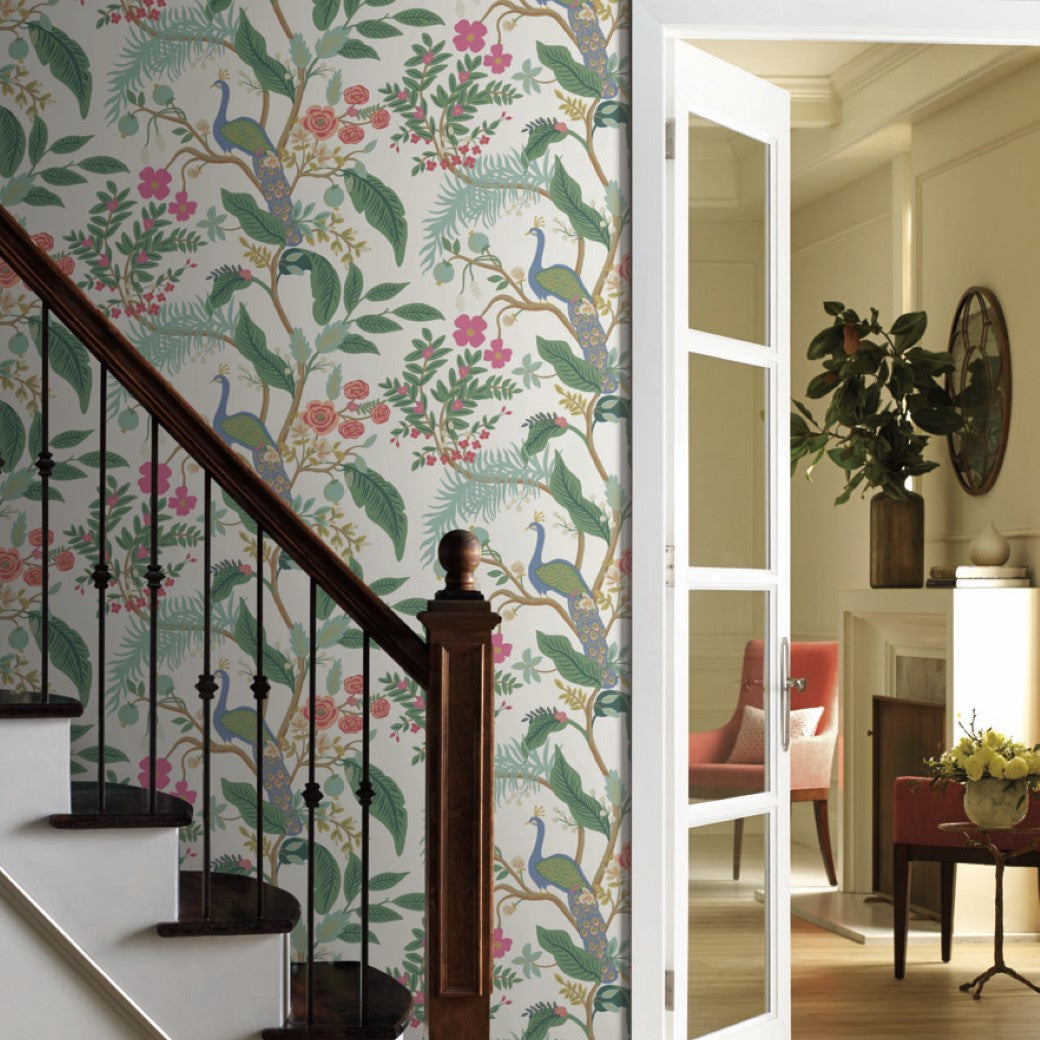 A staircase with a dark wooden banister is next to a wall adorned with vibrant York's Periwinkle Peacock Wallpaper, showcasing peacocks, flowers, and foliage. An open glass door leads to a cozy room with a fireplace and a potted plant.