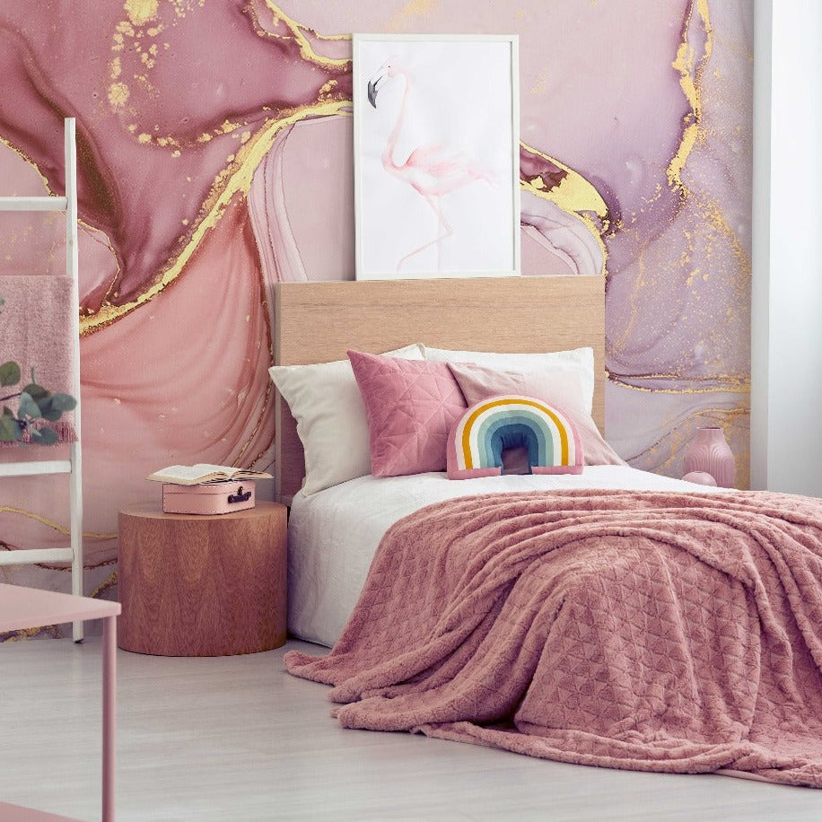 A stylish bedroom with a pink and gold theme, featuring a bed with a quilted blanket, a Decor2Go Wallpaper Mural, a rainbow-shaped pillow, and a small plant beside the bed.