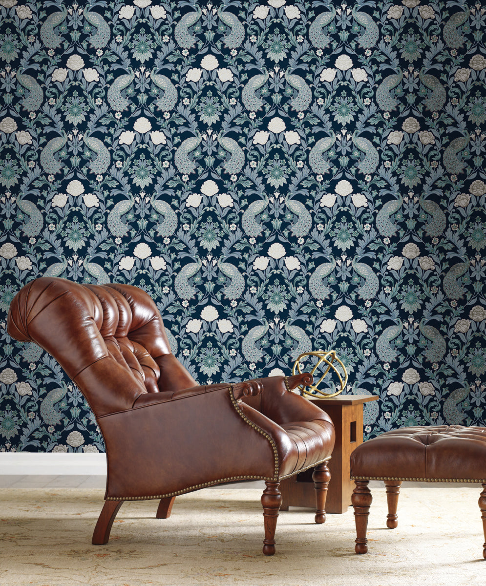 A luxurious brown leather armchair with a matching ottoman is placed beside a wooden side table holding a decorative metal object. The background features intricate Plume Dynasty Peacock & Floral Wallpaper - Navy (60 SqFt) by York Wallcoverings with stunning blue and white patterns, showcasing elegant floral and peacock motifs.