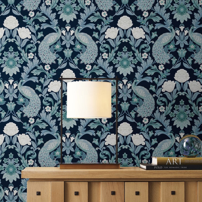 A table lamp sits on a wooden sideboard in front of an ornate dark blue and white Plume Dynasty Peacock & Floral Wallpaper - Navy (60 SqFt) from York Wallcoverings adorned with peacock motifs. Beside the lamp are two books, with one titled "ART," and a glass globe, adding a stylish touch to the decor.