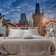A bed with white and grey bedding is set against a surreal backdrop of Prague Alley Wallpaper Mural by Decor2Go Wallpaper Mural, featuring iconic spires under a dramatic sunrise sky.