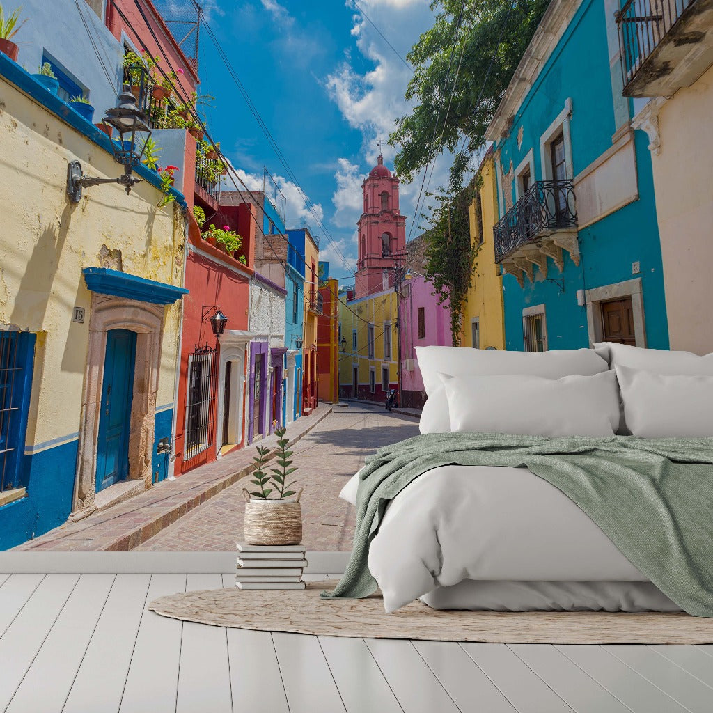 Bedroom - A surreal image blending a cozy bedroom setting with an outdoor vibrant street scene featuring colorful buildings and a clear blue sky. A bed is positioned in the foreground on white flooring, accented by Decor2Go Wallpaper Mural.