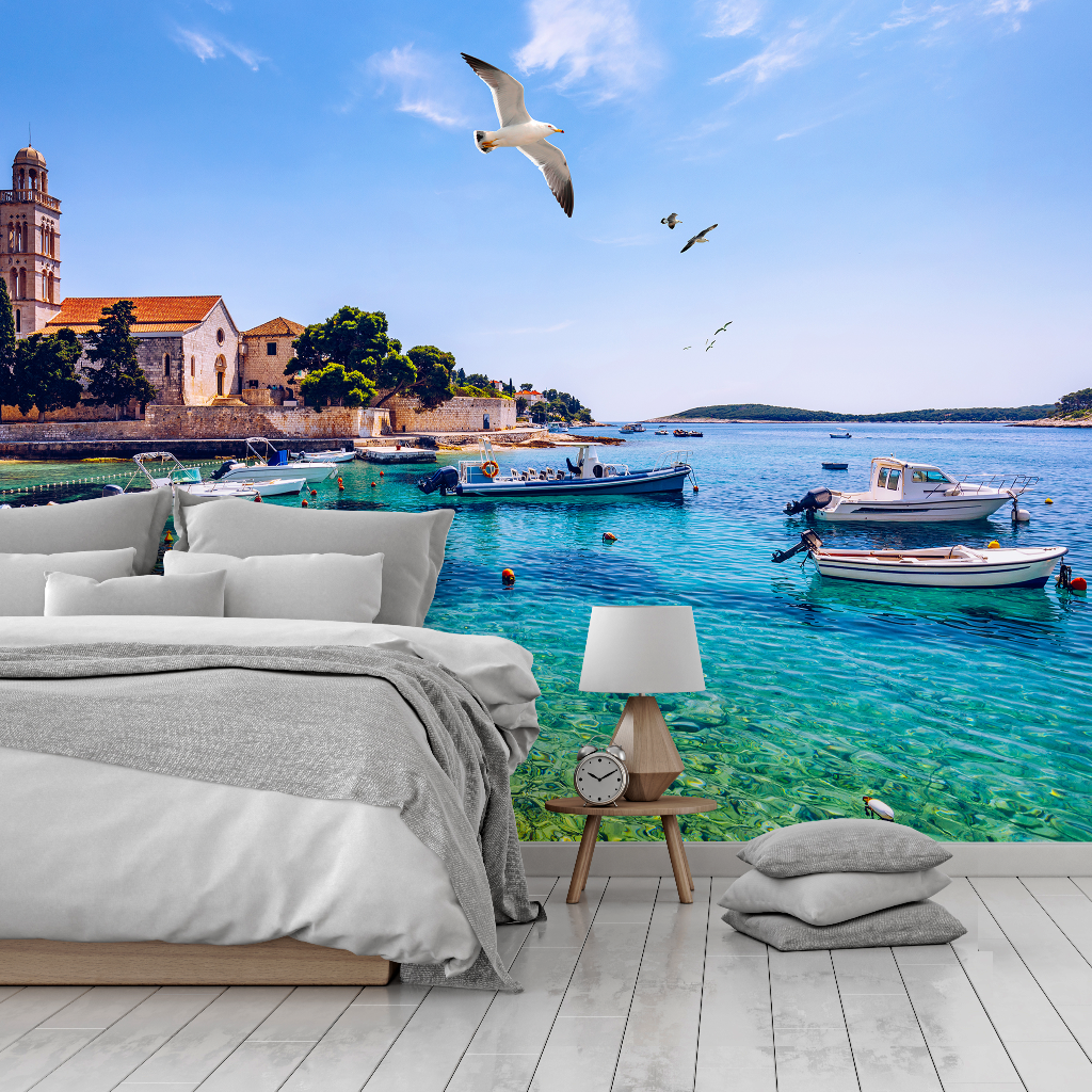 Bedroom - A bedroom with an immersive Decor2Go Wallpaper Mural of a scenic coastline featuring boats on clear blue water, a historic building in the background, and seagulls flying overhead.
