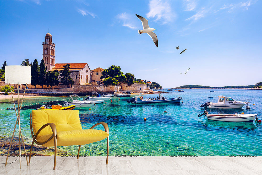 A scenic Italian Riviera view with clear blue waters, moored boats, and a historic building. 
In the foreground, a vibrant yellow chair faces the sea, and seagulls fly under a sunny Decor2Go Wallpaper Mural.