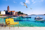 A scenic Italian Riviera view with clear blue waters, moored boats, and a historic building. 
In the foreground, a vibrant yellow chair faces the sea, and seagulls fly under a sunny Decor2Go Wallpaper Mural.