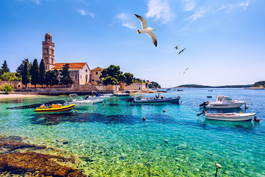 A picturesque Italian Decor2Go Wallpaper Mural coastal scene with clear turquoise waters, moored boats, a historic stone church with a bell tower, and flying seagulls under a bright blue sky.