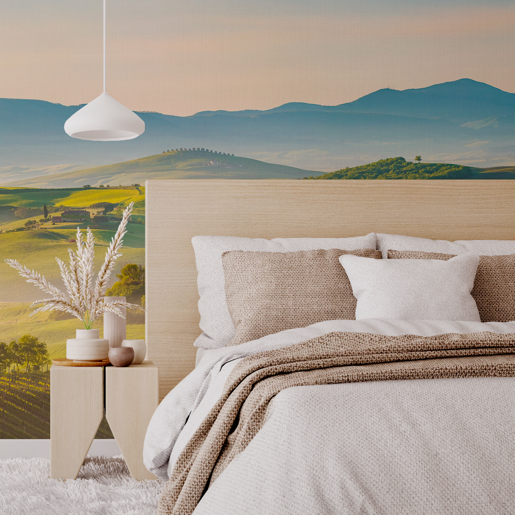 A cozy bedroom featuring a bed with neutral-toned bedding and multiple pillows, beside a wooden nightstand with decorative plants. A large Decor2Go Wallpaper Mural of Rolling Hills adds a tranquil backdrop.
