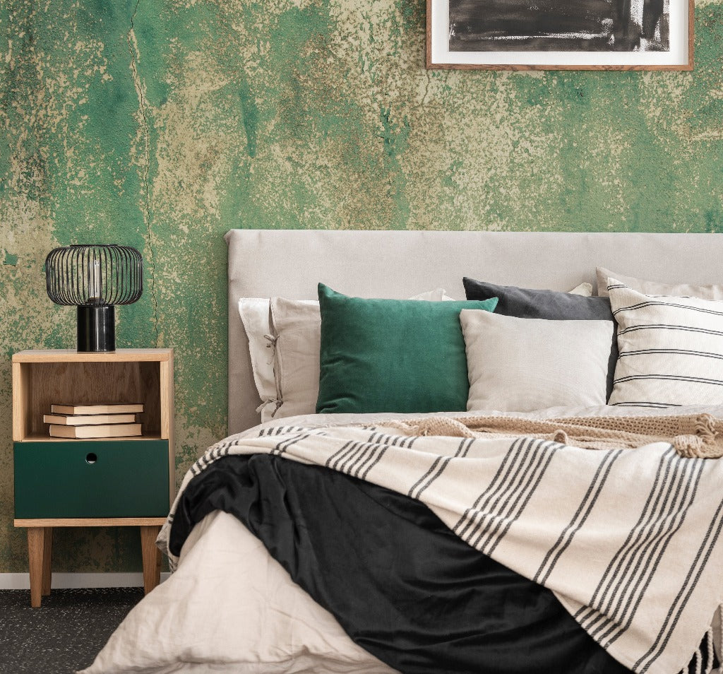 Bedroom - A stylish bedroom with a green textured accent wall, reminiscent of the Rusti rust Weathered Concrete mural wallpaper designs found in chic art studios from Decor2Go Wallpaper Mural. The room features a light gray upholstered bed and a variety of pillows in green, gray, and white. A wooden nightstand with a green drawer holds a modern wire-cage lamp beside the striped bedding.
