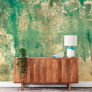 A wooden sideboard with a potted plant on the left and a white lamp with a blue base on the right. Two stacked books sit next to the lamp. The wall behind features a textured Decor2Go Wallpaper Mural Rusti rust Weathered Concrete mural wallpaper, while the floor is covered in grey carpet.