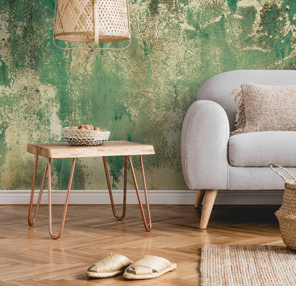 A small, rustic living space featuring a green textured Decor2Go Wallpaper Mural Rusti rust Weathered Concrete mural wallpaper, a light grey sofa with beige cushions, a wooden side table with hairpin legs holding a bowl, a woven pendant lamp, and a basket. Slippers and a beige rug are on the wooden floor. Perfect inspiration for design studios.