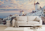 A modern living room with a white sofa and small tables in front of a large window displaying a Decor2Go Wallpaper Mural, showing its famous white houses and windmills against a sunset sky.