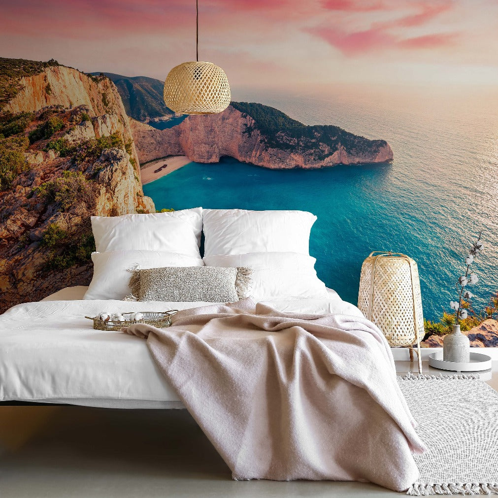 A luxurious bedroom with a large white bed and scenic ocean view through a floor-to-ceiling window, featuring cliffs and sunset skies. The decor includes hanging lights and Decor2Go Wallpaper Mural on the feature wall.