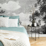 The cozy bedroom exudes timeless vintage elegance with a bed on the left, adorned with blue and white bedding. A crocheted blanket and an open book lie on the bed. A small round table holds candles and a vase with branches, set against the "Serene Forest Wallpaper Mural" by Decor2Go Wallpaper Mural, which showcases trees and clouds.