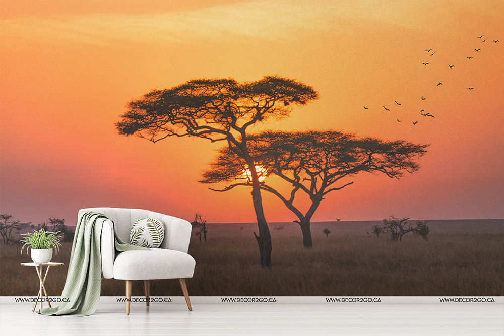 A sunset in a savannah-themed bedroom decor with a Decor2Go Wallpaper Mural displaying an orange sky, silhouetted acacia trees, and flying birds. A white armchair, throw