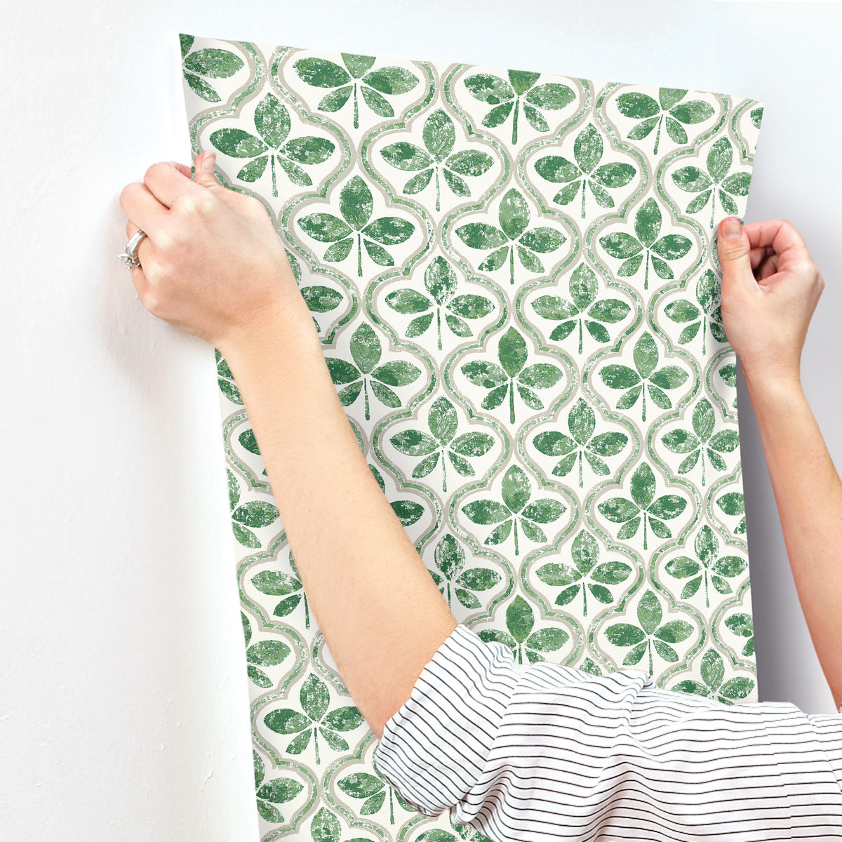 A person is applying York Wallcoverings Sevilla Ochre Wallpaper Yellow (60 Sq.Ft.) with a green and white clover pattern to a wall. The person's hands are smoothing the wallpaper into place, and they are wearing a striped shirt with rolled-up sleeves and a ring on one finger, showcasing geometric elegance in every detail.