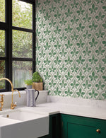 A kitchen corner featuring a green leaf pattern backsplash, complemented by Sevilla Ochre Wallpaper Yellow (60 Sq.Ft.) by York Wallcoverings for geometric elegance. It boasts a white marble countertop and dark green cabinets. A white farmhouse sink with a brass faucet is beneath black-framed windows. Accessories include a small plant, a striped pitcher, and a gold cabinet handle.