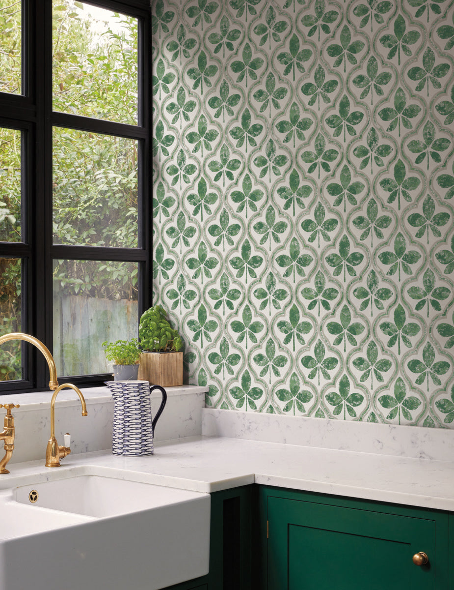 A kitchen corner featuring a green leaf pattern backsplash, complemented by Sevilla Ochre Wallpaper Yellow (60 Sq.Ft.) by York Wallcoverings for geometric elegance. It boasts a white marble countertop and dark green cabinets. A white farmhouse sink with a brass faucet is beneath black-framed windows. Accessories include a small plant, a striped pitcher, and a gold cabinet handle.