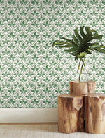 A vase with a large green leaf sits on two stacked wooden stools in a room where York Wallcoverings Sevilla Onyx Wallpaper Black, Brown (60 Sq.Ft.) featuring green and white floral designs brings an extra touch of charm. The scene is complemented by a light-colored carpet on the floor, blending natural beauty with geometric elegance.