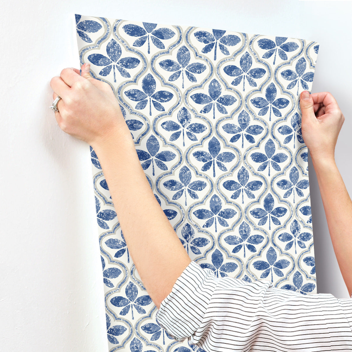 Person with a striped shirt and a ring on their finger smoothing out a section of Sevilla Ochre Wallpaper Yellow (60 Sq.Ft.) by York Wallcoverings with a blue and white clover-like pattern on a white wall.