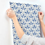 A person with rolled-up striped sleeves is applying blue and white patterned Sevilla Cobalt Wallpaper Blue (60 Sq.Ft.) by York Wallcoverings with a repeating clover motif to a wall. Their hands hold the wallpaper at the top, ensuring it aligns correctly. The background wall is plain white, highlighting its geometric elegance.