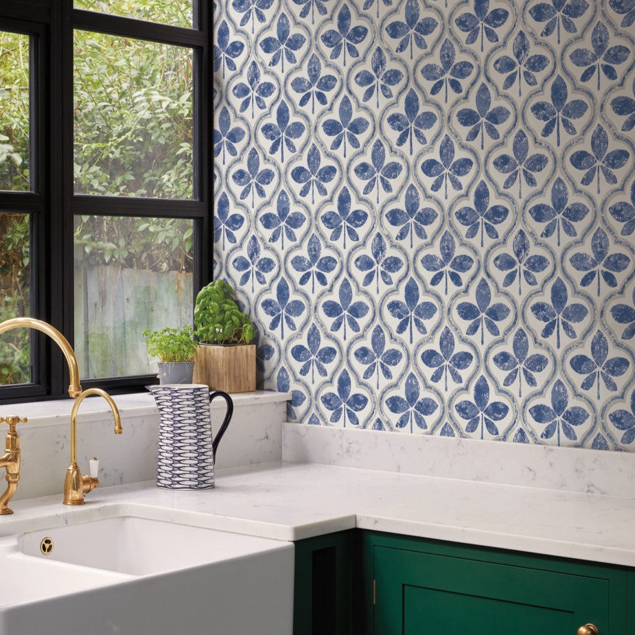 A kitchen corner with a green cabinet, white marble countertops, and a white farmhouse sink. The backsplash features Sevilla Ochre Wallpaper Yellow (60 Sq.Ft.) by York Wallcoverings. There's a potted plant and a blue patterned jug on the counter. Black-framed windows overlook greenery, complementing the geometric elegance of the space.