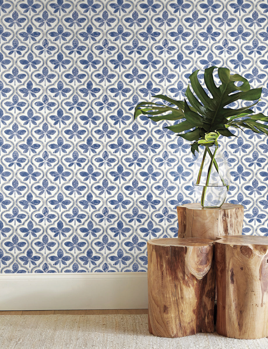 A room with York Wallcoverings Sevilla Cobalt Wallpaper Blue (60 Sq.Ft.) featuring a blue and white floral pattern. A clear vase holding large, green monstera leaves is placed on a wooden stump table. The table sits on a beige carpet, and the scene exudes minimalist geometric elegance.