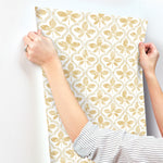 Person with a striped shirt holding and positioning Sevilla Ochre Wallpaper Yellow (60 Sq.Ft.) featuring a gold butterfly design against a white wall. The person, wearing a ring on the left hand, carefully aligns the York Wallcoverings wallpaper for seamless application.