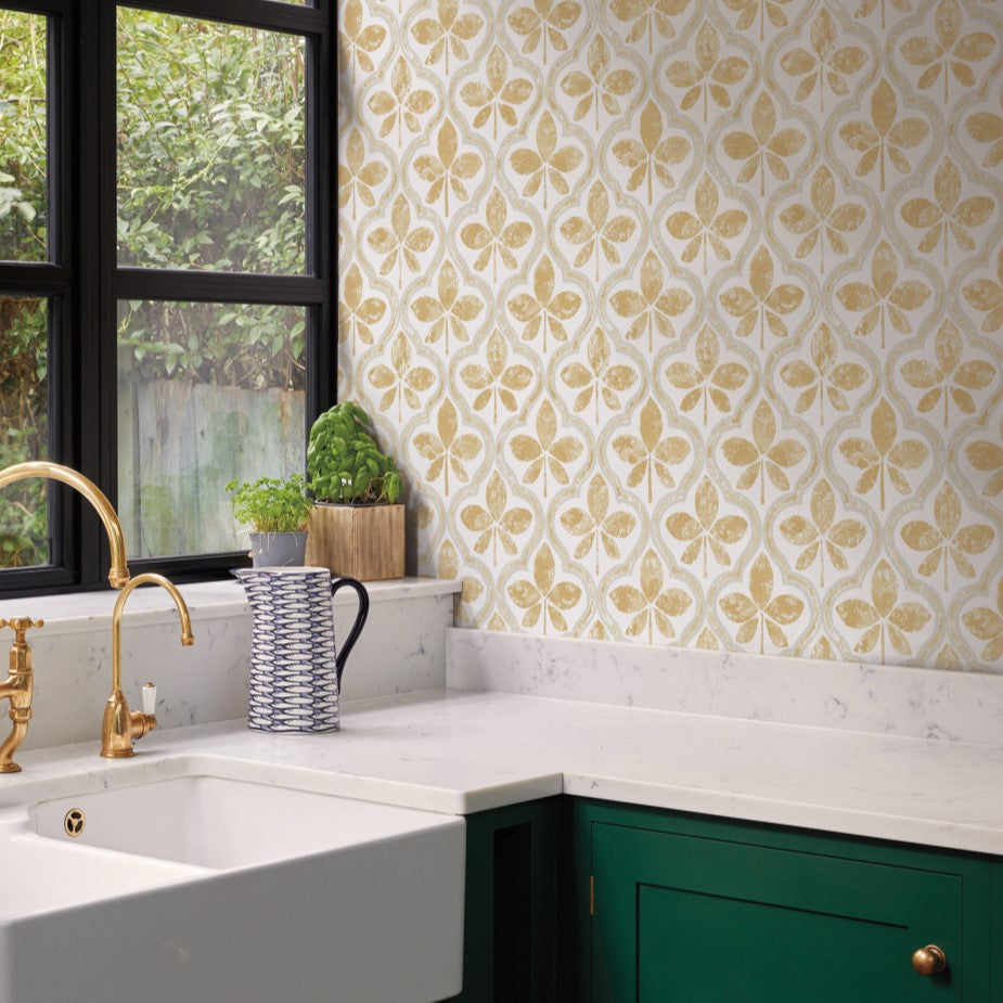 A kitchen with white marble countertops, green cabinets, and brass fixtures. A large patterned Sevilla Cobalt Wallpaper Blue (60 Sq.Ft.) by York Wallcoverings featuring golden leaf designs adorns the wall, adding geometric elegance. A potted plant and a striped mug sit on the counter near a black-framed window with a view of greenery.