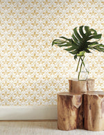 A minimalist interior features York Wallcoverings' Sevilla Ochre Wallpaper Yellow (60 Sq.Ft.) showcasing geometric elegance in yellow and white. A wooden side table with a natural cut hosts a glass vase containing large green leaves. The room has a light-colored carpet and a simple, clean, and natural aesthetic.