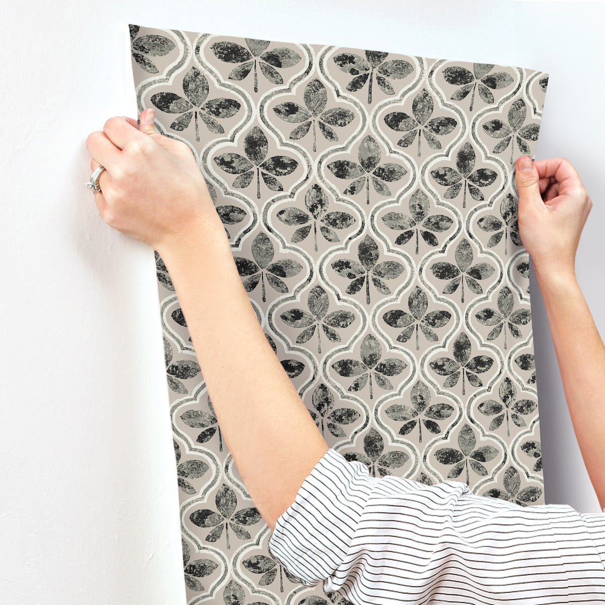 A person with a ring on their left hand is applying York Wallcoverings Sevilla Cobalt Wallpaper Blue (60 Sq.Ft.) featuring a black and white patterned design of butterflies and clover-like shapes onto a white wall. The person is wearing a long-sleeved, striped shirt, showcasing geometric elegance in their decor choice.