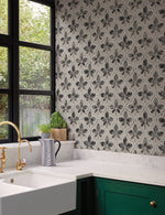 A kitchen corner with a green cupboard, white countertop, and white farmhouse sink. The wall showcases **Sevilla Ochre Wallpaper Yellow (60 Sq.Ft.) from York Wallcoverings** with a black and white patterned tile backsplash, and there's a large window with a view of greenery outside. On the countertop, there's a striped pitcher and a small potted plant.