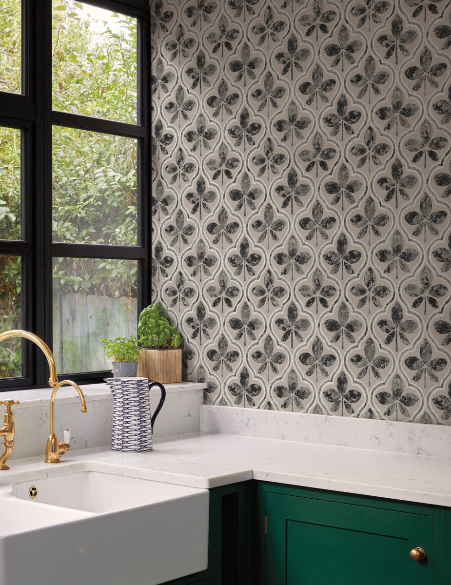 A bright kitchen corner features an ornate black and white tiled backsplash, large black-framed windows, and a white farmhouse sink with gold faucets. Green cabinetry and a white countertop with a blue and white striped jug, a potted plant, and York Wallcoverings' Sevilla River Rock Wallpaper Grey (60 Sq.Ft.) add to the Geometric Elegance of the decor.